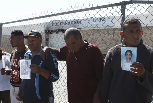 Migrants, mostly from Venezuela, hold photos of those who died in a fire at a Mexican immigration detention center, behind, during a prayer vigil outside the center in Ciudad Juarez, Mexico, April, 27, 2023. Four months after a fire in a Mexican immigration detention center at the border killed 40 migrants, some survivors are living in limbo at a Mexico City hotel, recovering from their injuries and awaiting the prosecution of their captors. (AP Photo/Christian Chavez, File)