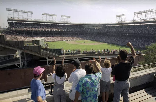 In this Monday, July 9, 1990, file photo, spectators watch an All-Star Game practice session from the roof of a building just outside Chicago's Wrigley Field. Booking hotels for baseball games during shoulder seasons like May, June or September can often be cheaper than the summer months. Additionally, save money by planning to attend weekday versus weekend games. (AP Photo/Seth Perlman, File)