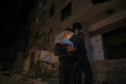 Two boys look at a cellphone in a dark street in Damascus, Syria, early Thursday March 27, 2025. Neighborhoods in most parts of the country turn pitch black once the sunsets, save on some lights from street lamps, Mosque minarets, and drivers with their floodlights on to see.(AP Photo/Omar Sanadiki)