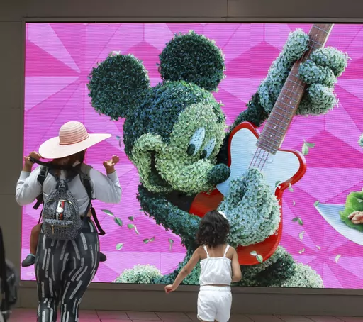 Travelers check out a jumbo screen projecting images of Walt Disney World in the east hall atrium at Orlando International Airport, Thursday, May 25, 2023 in Orlando, Fla. Auto club AAA said this summer could be “one for the record books, especially at airports,” with more than 43 million Americans projected to travel 50 miles or more. (Joe Burbank /Orlando Sentinel via AP)